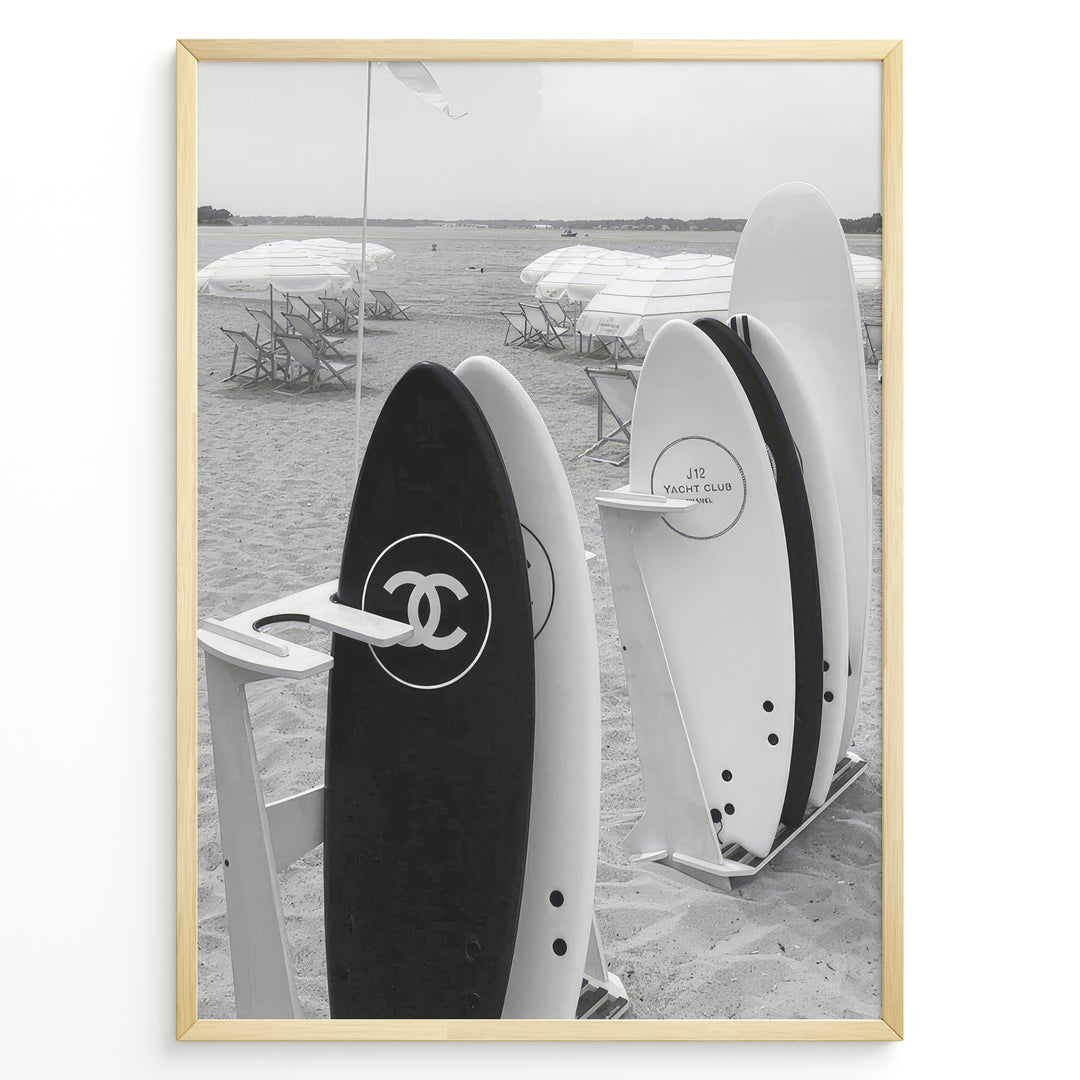 Fashion Plakat | Chanel Surfboards on the Beach – Luxury Meets Coastal Lifestyle in Black and White Plakaty.dk | Black and white photo of Chanel and J12 Yacht Club surfboards on a sandy beach with umbrellas and lounge chairs in the background