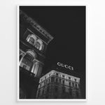 Ladda upp bild till gallerivisning, Fashion Plakat | Night View of Gucci Building Facade Plakaty.dk | Black and white nighttime photo of an ornate building with classical columns and arches next to a building displaying a large illuminated &quot;GUCCI&quot; sign against a dark sky.
