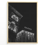 Ladda upp bild till gallerivisning, Fashion Plakat | Night View of Gucci Building Facade Plakaty.dk | Black and white nighttime photo of an ornate building with classical columns and arches next to a building displaying a large illuminated &quot;GUCCI&quot; sign against a dark sky.
