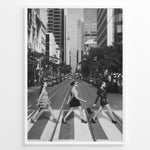 Ladda upp bild till gallerivisning, Fashion Plakat | Three Women on a Crosswalk in a City Plakaty.dk | Et indrammet sort-hvidt fotografi, der viser tre kvinder, der går fra venstre mod højre på et fodgængerfelt i en by. Kvinden til venstre har en kjole på, kvinden i midten har en top og nederdel, og kvinden til højre har en kort, mørk kjole på og bærer en taske. Høje skyskrabere og et tårn er synlige i baggrunden, med biler parkeret langs gaden.
