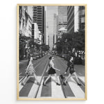 Ladda upp bild till gallerivisning, Fashion Plakat | Three Women on a Crosswalk in a City Plakaty.dk | Et indrammet sort-hvidt fotografi, der viser tre kvinder, der går fra venstre mod højre på et fodgængerfelt i en by. Kvinden til venstre har en kjole på, kvinden i midten har en top og nederdel, og kvinden til højre har en kort, mørk kjole på og bærer en taske. Høje skyskrabere og et tårn er synlige i baggrunden, med biler parkeret langs gaden.

