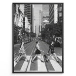 Ladda upp bild till gallerivisning, Fashion Plakat | Three Women on a Crosswalk in a City Plakaty.dk | Et indrammet sort-hvidt fotografi, der viser tre kvinder, der går fra venstre mod højre på et fodgængerfelt i en by. Kvinden til venstre har en kjole på, kvinden i midten har en top og nederdel, og kvinden til højre har en kort, mørk kjole på og bærer en taske. Høje skyskrabere og et tårn er synlige i baggrunden, med biler parkeret langs gaden.
